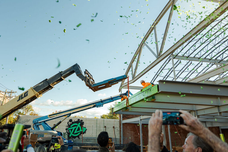 Construction workers secure a steel beam at the future Trueline entertainment venue in Greenville as green confetti falls and people watch and take photos. The milestone is part of Brasfield & Gorrie’s ongoing work on the site.