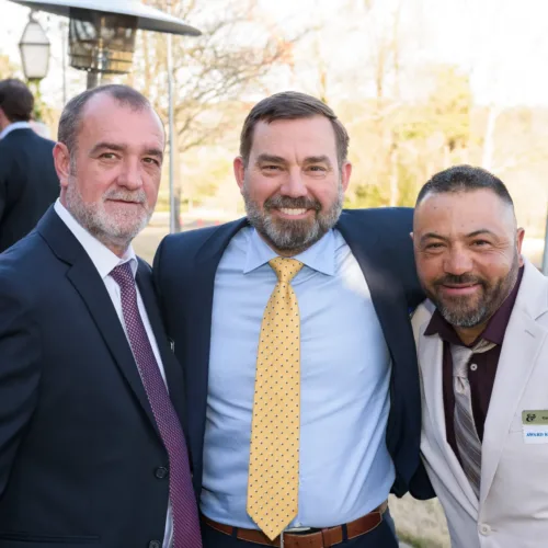 Three men in suits, including James Cohran, pose together outdoors at a formal event, smiling for the camera. One man wears a name tag amid trees and patio heaters in the background.