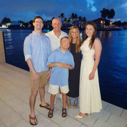 Five people, including Dan Spinetto, stand together on a waterfront promenade at dusk, with palm trees, water, and houses visible in the background.