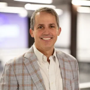 A man in a light plaid blazer and collared shirt smiles while standing indoors with a blurred office background.