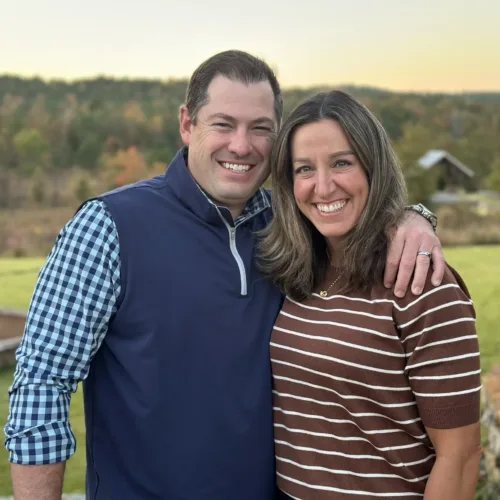 A man and woman stand outdoors, smiling at the camera, with trees and a grassy field in the background. The man has his arm around the woman's shoulder.