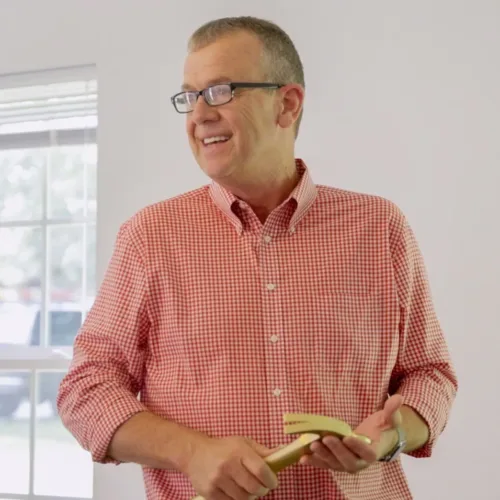 Kenneth Underwood, wearing glasses and a red checkered shirt, stands indoors holding a golden hammer in front of a mirror.