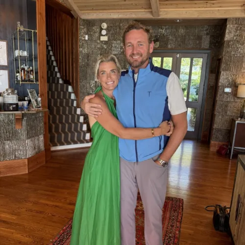 A woman in a green dress and a man in a blue vest stand smiling and hugging on a patterned rug in a wood-paneled room.
