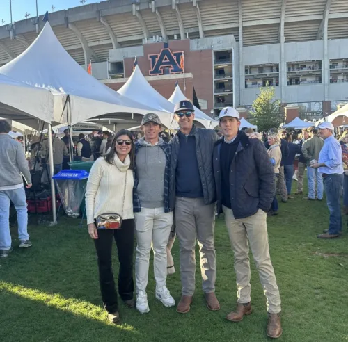 Four people, including Seth Sargent, stand and pose for a photo in front of Auburn University’s football stadium during a tailgate event. Tents and other attendees are visible in the background.