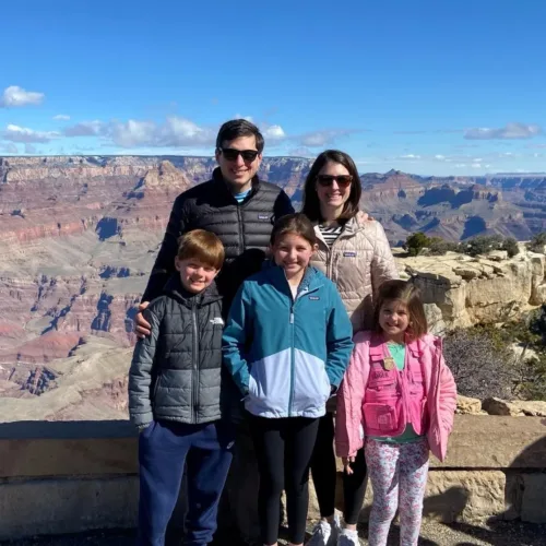 A family of five poses in jackets and casual clothes at a canyon overlook on a sunny day with clear blue sky and canyon views in the background.