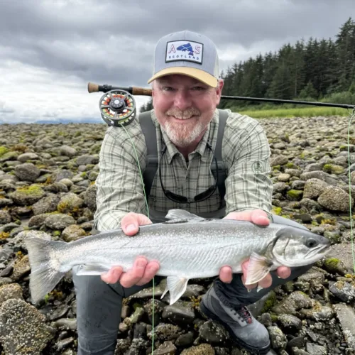 Chris Britton, dressed in outdoor clothing, kneels on rocky ground, holding a large fish with both hands and a fly fishing rod over his shoulder.