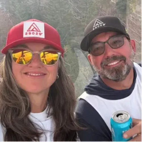 Two people wearing hats and sunglasses smile at the camera; one holds a blue can. Trees and sky are visible in the background.