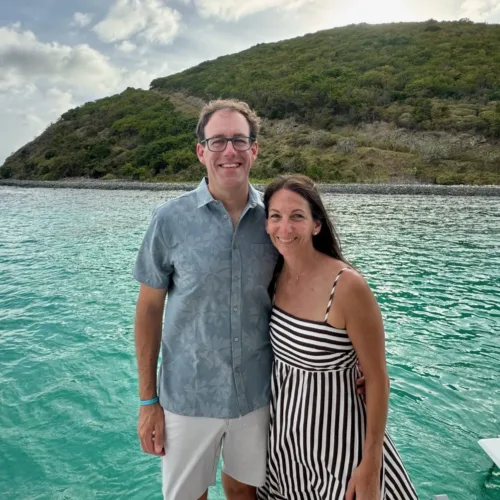Greg Hunsberger and a woman stand together on a boat near clear turquoise water, with a green hillside in the background under a partly cloudy sky.