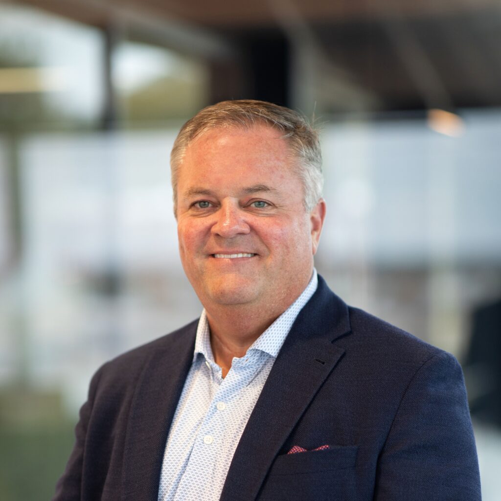 Reed Nelson, a middle-aged man in a navy suit jacket and light shirt, smiles as he stands indoors with blurred glass windows in the background.