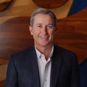Eric Perkinson, dressed in a dark suit jacket and white shirt, stands in front of a wooden wall with abstract patterns, smiling at the camera.