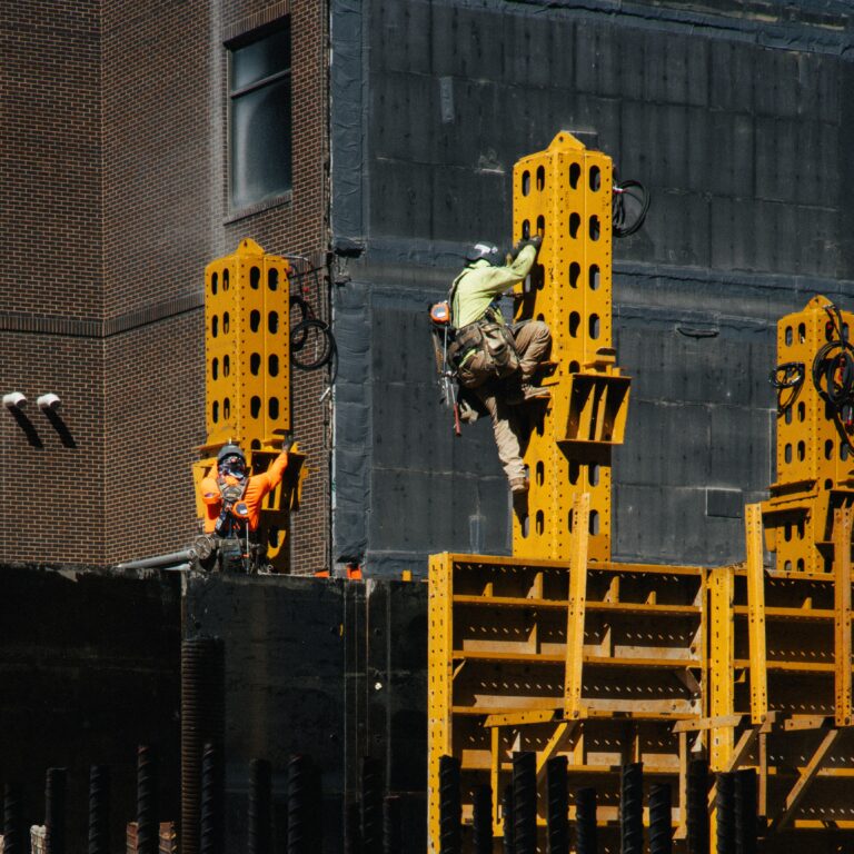 A construction worker in safety gear climbs a yellow steel beam structure at a building site, demonstrating the 7 commitments, while another worker stands nearby.