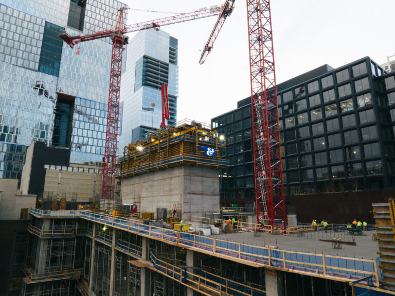 A construction site with cranes, scaffolding, and workers from Brasfield & Gorrie, surrounded by modern glass office buildings in an urban setting.