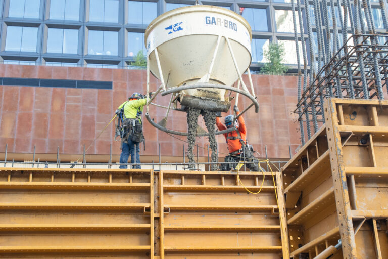 Two trades workers guide wet concrete pouring from a large funnel into a metal formwork at a building site.