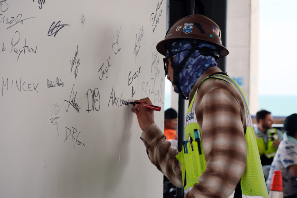 A construction worker in safety gear showcases craftsmanship as he signs a white wall already covered with various signatures and markings.