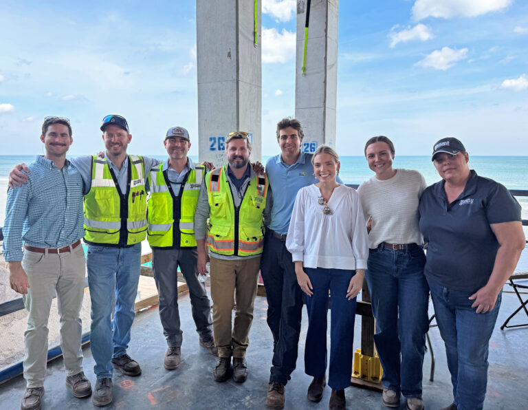 Eight people, some in safety vests and hard hats, stand together on a construction site near the ocean, smiling at the camera.