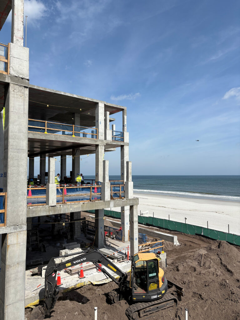 A multi-story concrete building under construction near the coastline, with workers and construction equipment, including an excavator, showcasing skilled craftsmanship in the foreground.