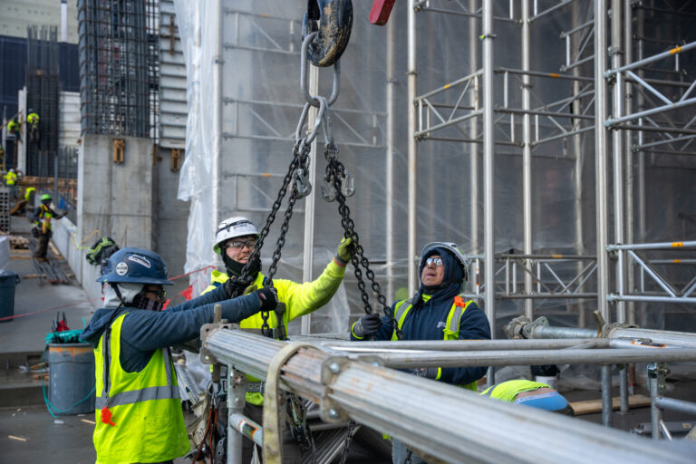 Construction workers in safety gear use chains and a crane to lift metal pipes at a Brasfield and Gorrie Georgia careers building site, with scaffolding and protective coverings in the background.