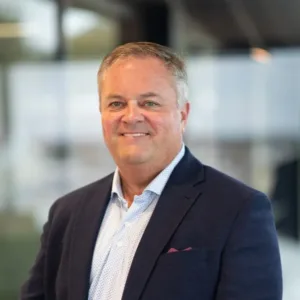 Man in a navy suit and light blue shirt, standing indoors with blurred windows and office background, smiling at the camera.