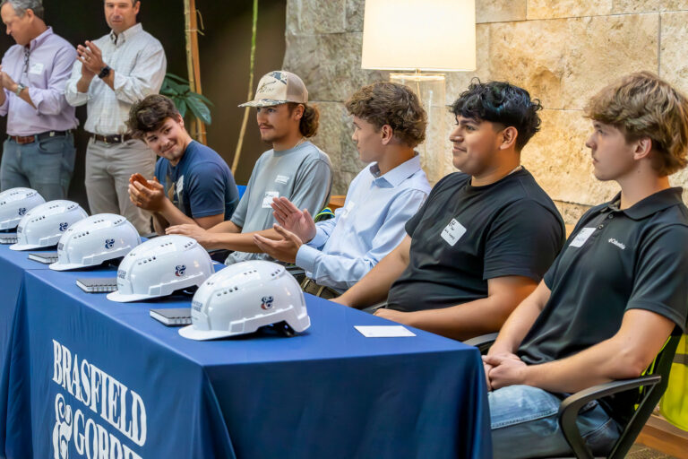 Five young men sit behind a table with hard hats and a 