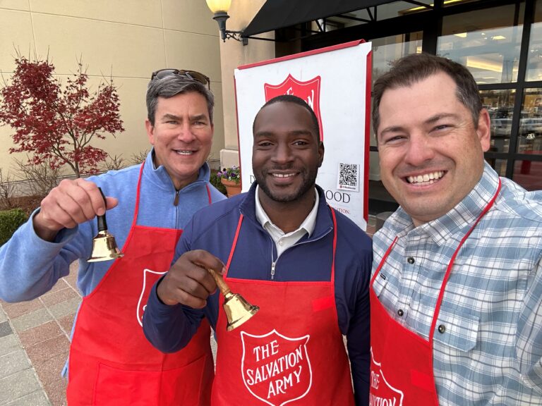 Three men wearing red Salvation Army aprons and holding bells smile for a photo outside a building with a Salvation Army sign, spreading cheer and encouraging charity donations.