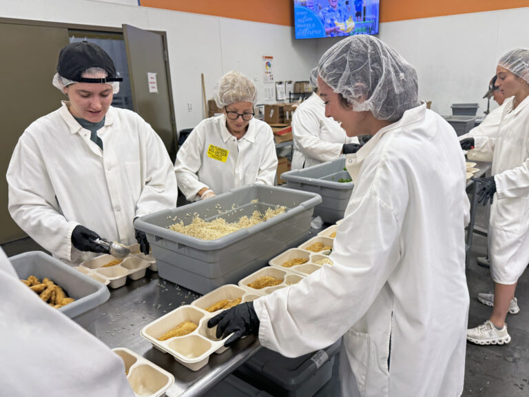Several people in white lab coats, hairnets, and gloves are assembling meal trays at a worktable in a commercial kitchen, supporting charity through the preparation of meals for those in need.