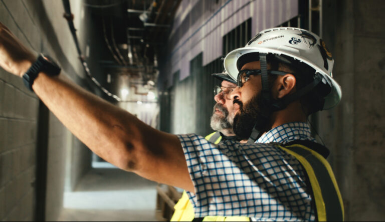 Dos obreros de la construcción, equipados con cascos y chalecos reflectantes, inspeccionan o comentan algo sobre una pared en el interior de un edificio sin terminar, en un proyecto de Brasfield & Gorrie.