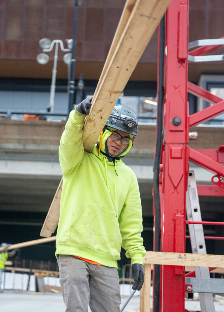 Un obrero de la construcción, vestido con una sudadera amarilla con capucha y un casco de seguridad, levanta una larga viga de madera en una obra al aire libre de Brasfield & Gorrie; al fondo se ven otros trabajadores y maquinaria.
