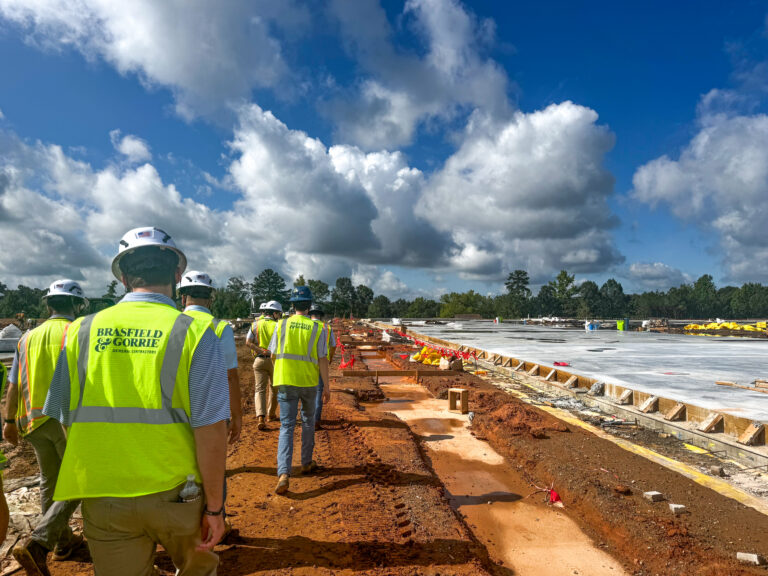 Un grupo de trabajadores en prácticas y en programas de formación dual de Brasfield and Gorrie, equipados con chalecos de seguridad y cascos, camina por un camino de tierra en una obra al aire libre bajo un cielo parcialmente nublado.