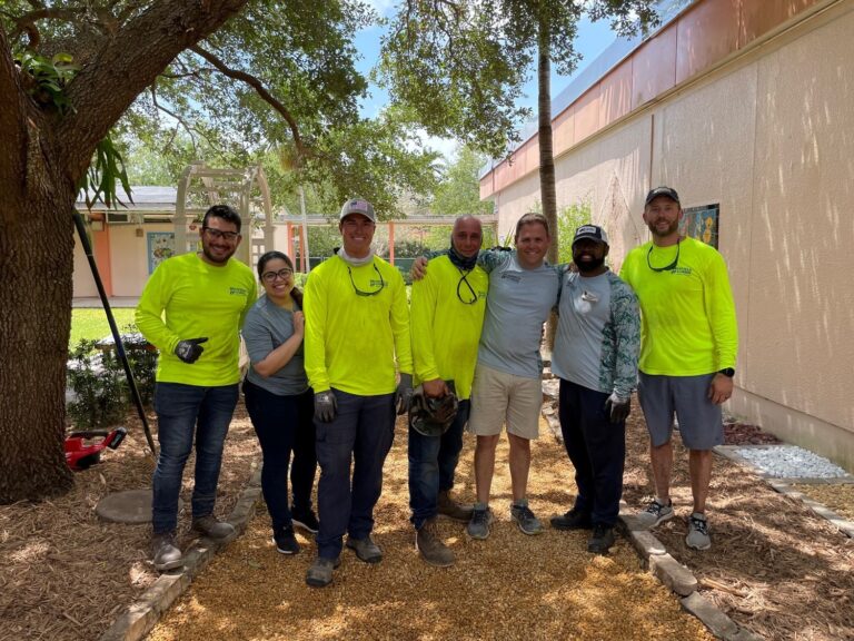Seven people stand on a gravel path outdoors, some wearing neon yellow shirts and work gloves, showcasing their community involvement as they pose for a group photo near a building and a tree.