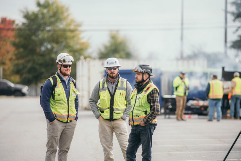 Tres obreros de la construcción, ataviados con chalecos de seguridad y cascos, posan juntos en un aparcamiento, en representación de Brasfield & Gorrie, con otros trabajadores y maquinaria visibles al fondo.