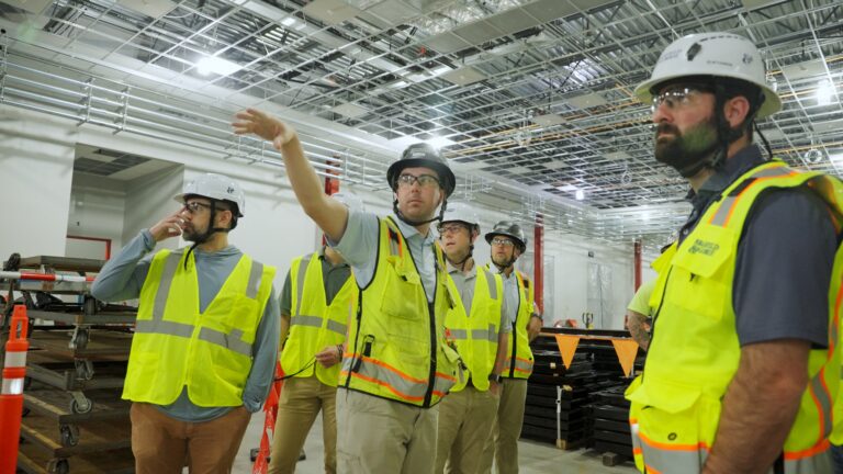 Varios obreros de la construcción, equipados con cascos y chalecos reflectantes, se encuentran en el interior del edificio; uno de ellos gesticula y señala, mientras los demás observan el techo sin terminar: una muestra de la innovación de Brasfield and Gorrie impulsada por la tecnología VDC.