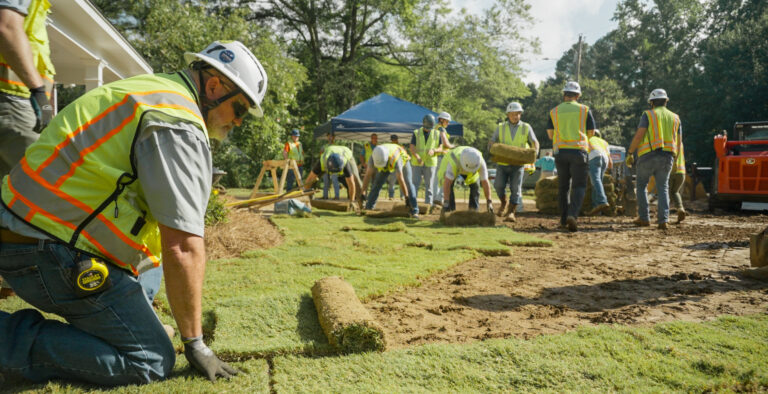 Workers in safety vests and helmets lay sod on a lawn, showcasing community involvement as more people and equipment are visible in the background on a sunny day.