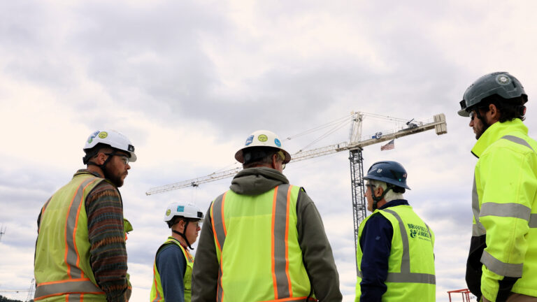 Five Brasfield & Gorrie construction workers in safety gear and helmets stand on a site with a crane and cloudy sky in the background.