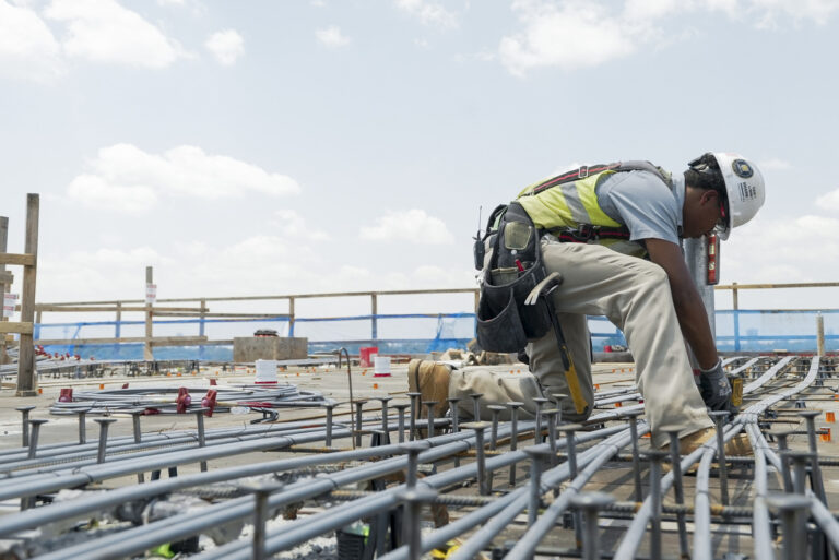 Construction worker wearing safety gear installing electrical conduit pipes on a building site under a clear sky, showcasing the hands-on opportunities available with Brasfield and Gorrie careers.