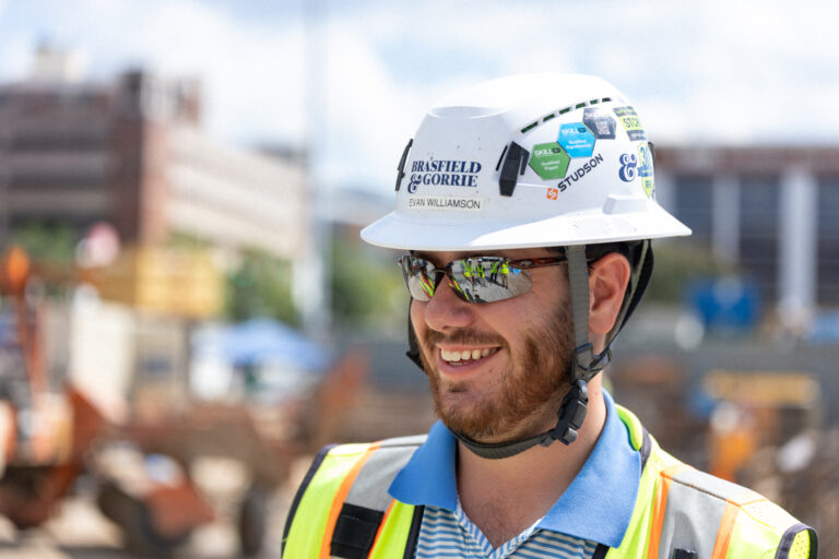 A man wearing a hard hat, safety vest, and sunglasses smiles at a construction site, showcasing the rewarding opportunities offered by Brasfield and Gorrie careers.