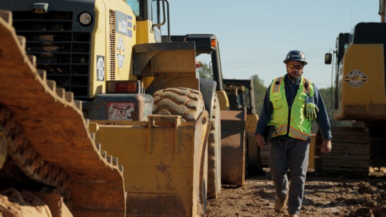 Un obrero de la construcción, con chaleco de seguridad y casco, camina junto a una gran máquina de obra amarilla en una obra sin asfaltar de Brasfield & Gorrie.