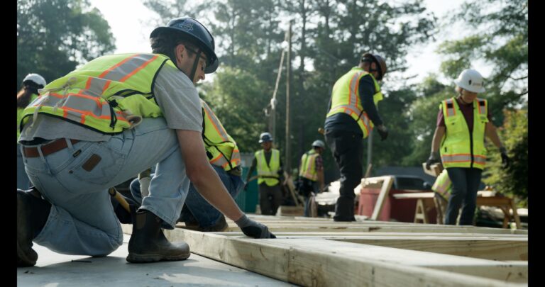 Construction workers wearing safety vests and helmets assemble wooden frames outdoors on a building site surrounded by trees, illustrating the hands-on teamwork found in Brasfield and Gorrie careers.