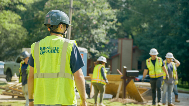 A construction worker wearing a Brasfield & Gorrie safety vest and helmet stands outdoors, while several workers—including participants in Brasfield and Gorrie student intern co-op programs—are active at the building site in the background.