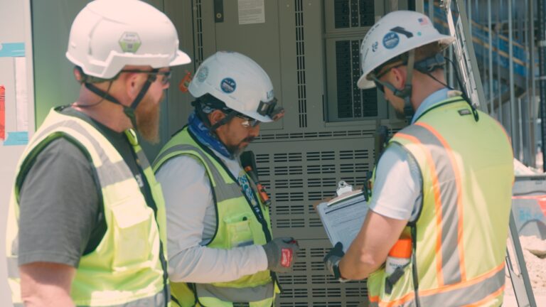 Tres trabajadores de la constructora Brasfield & Gorrie, equipados con material de seguridad, revisan documentos y equipos en una obra al aire libre.