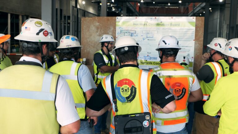 A group of construction workers in safety vests and helmets, including Brasfield and Gorrie student intern co-op program participants, gather around a coordination board at a worksite.