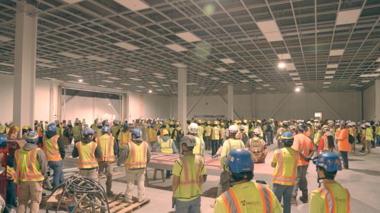 Large group of construction workers in safety vests and helmets gathered inside a spacious unfinished building for a meeting or briefing about construction services careers.