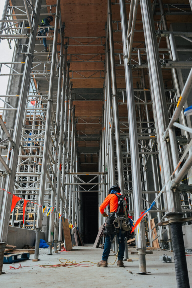 A construction worker in safety gear stands among metal scaffolding at a Brasfield & Gorrie building site while another worker is elevated above on the scaffolding.