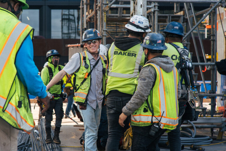 Un grupo de obreros de la construcción, equipados con material de seguridad y cascos, se reúne en una obra muy concurrida, llena de andamios y materiales, lo que refleja la ventaja competitiva de Brasfield and Gorrie a la hora de ejecutar proyectos complejos de forma segura.