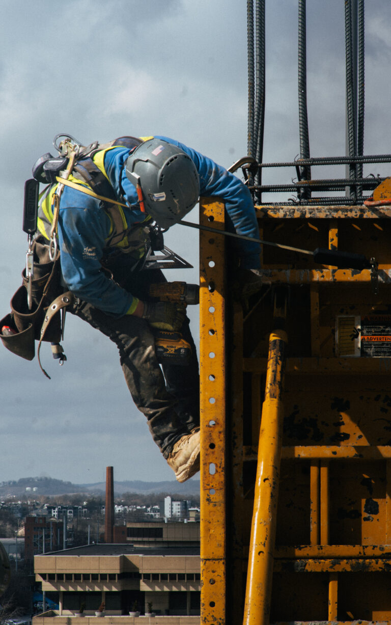 A Brasfield & Gorrie construction worker in safety gear and helmet uses a power tool while working on a steel structure high above a cityscape.