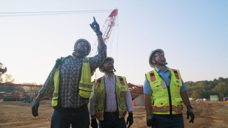 Three construction workers in safety gear and helmets look up at a construction site; one points toward something in the distance, with a crane visible in the background—showcasing opportunities with Brasfield and Gorrie careers.