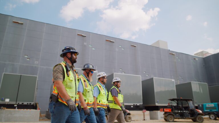 Four construction workers wearing safety gear walk in front of a large industrial building on a sunny day, showcasing the teamwork and opportunity found in Brasfield and Gorrie careers.