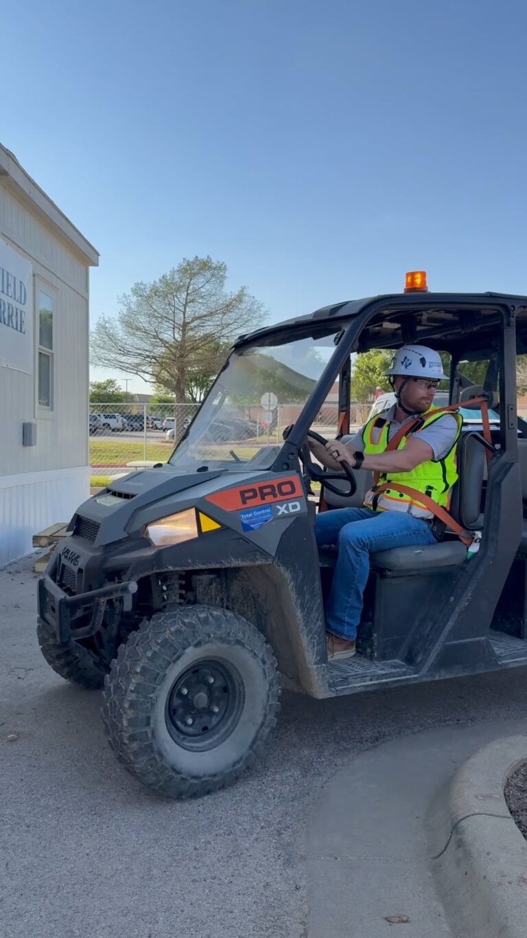 A construction worker in a safety vest and hard hat sits in a utility vehicle with a flashing orange light near a portable building on a sunny day, representing the rewarding opportunities available through Brasfield and Gorrie careers.
