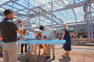 A group gathers at the construction site for the Christ Church Episcopal School expansion, with some signing a blue steel beam and another person recording the event. Brasfield & Gorrie’s structural framework is visible in the background.
