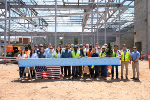 A group in safety gear poses at the Christ Church Episcopal School construction site behind a signed steel beam with a small tree and U.S. flag, celebrating the school expansion by Brasfield & Gorrie. Structural framework rises in the background.