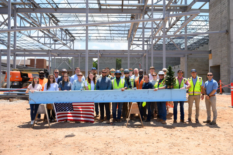 A group in safety gear poses at the Christ Church Episcopal School construction site behind a signed steel beam with a small tree and U.S. flag, celebrating the school expansion by Brasfield & Gorrie. Structural framework rises in the background.
